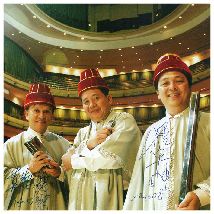 Trois harmonicistes en costume traditionnel dans une salle de concert, photo dédicacée